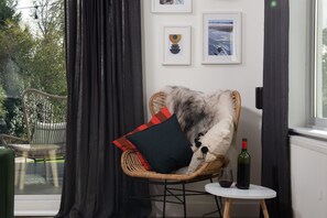 A wicker chair with a pillow, a small table with a bottle and glass, and framed pictures on the wall. - Meadow Retreat - Sleeps 12 | 4 Bedrooms | Hot Tub | Sauna | Cinema Room | Games Room | Dog Friendly (Hatt, Saltash, Cornwall)