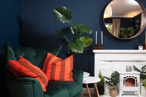 A room with a green velvet armchair, a round mirror, and a white fireplace. - Meadow Retreat - Sleeps 12 | 4 Bedrooms | Hot Tub | Sauna | Cinema Room | Games Room | Dog Friendly (Hatt, Saltash, Cornwall)