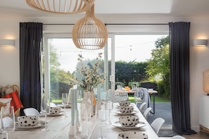 A dining area with a long table set for a meal, a vase with flowers, and a view of a garden through large windows. - Meadow Retreat - Sleeps 12 | 4 Bedrooms | Hot Tub | Sauna | Cinema Room | Games Room | Dog Friendly (Hatt, Saltash, Cornwall)