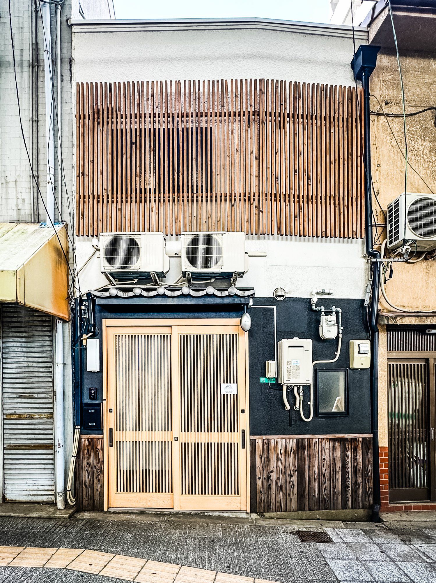 Fachada de edificio con pared de listones de madera, puerta metálica y unidades de aire acondicionado.