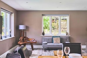A living room with a sofa, a dining table with chairs, a lamp, and a window with a view of the outside. - Heronbrook Retreat, Bedfordshire | Outdoor Pool, Hot Tub, Sleeps 20 (Bedford)