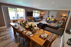 A dining area with a long wooden table set for a meal, a sofa, and a television in the background. - Heronbrook Retreat, Bedfordshire | Outdoor Pool, Hot Tub, Sleeps 20 (Bedford)