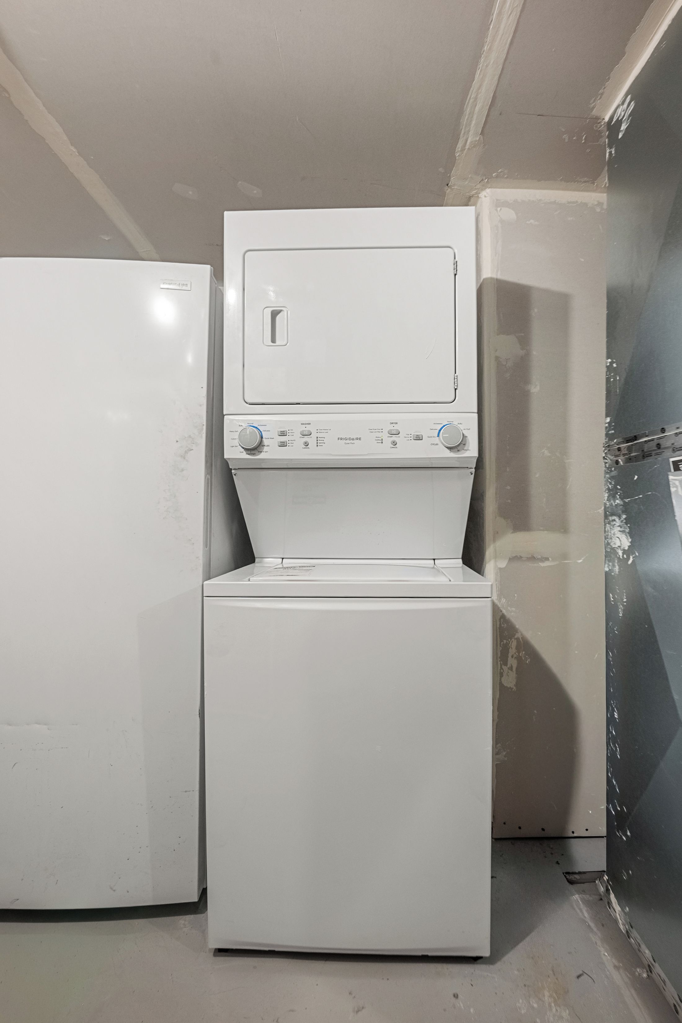 A laundry room with a stacked washer and dryer, a white refrigerator, and a wall with peeling paint.