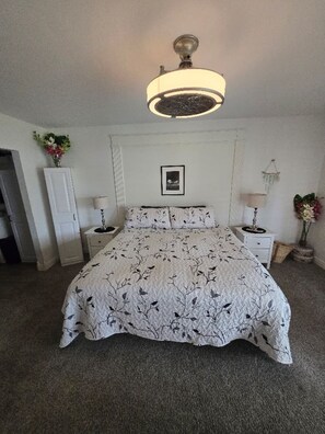 Ein Schlafzimmer mit einem Bett, zwei Nachttischen, einer Stehlampe, einer Deckenleuchte, einem gerahmten Bild an der Wand und einer Tür.