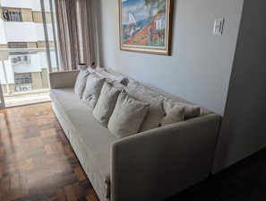 A modern living room with a sofa, a painting on the wall, and a view of the outside. - Apartment one block from the beach, central location, close to many attractions. (Balneário Camboriú)