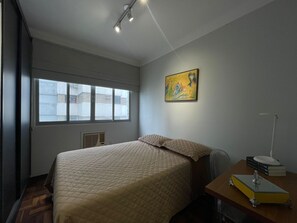 A bedroom with a bed, a desk with a lamp, a painting, and a window with a view of buildings. - Apartment one block from the beach, central location, close to many attractions. (Balneário Camboriú)