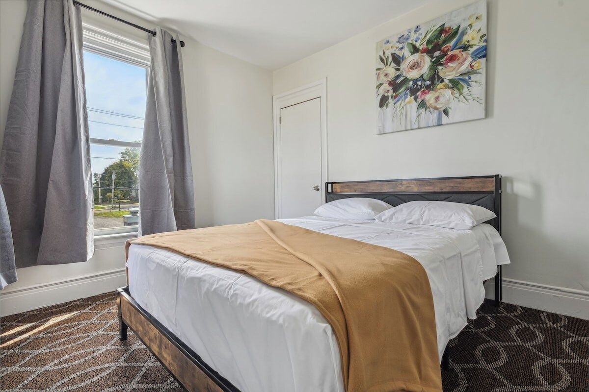 A bedroom with a bed, a window with curtains, a door, and a floral painting on the wall.