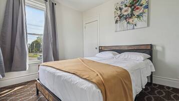 A bedroom with a bed, a window with curtains, a door, and a floral painting on the wall.