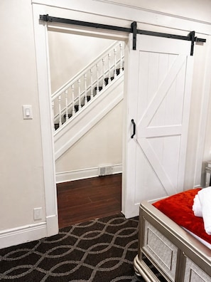 Un dormitorio con una cama, un plaid rojo, una puerta blanca, una escalera y un radiador.