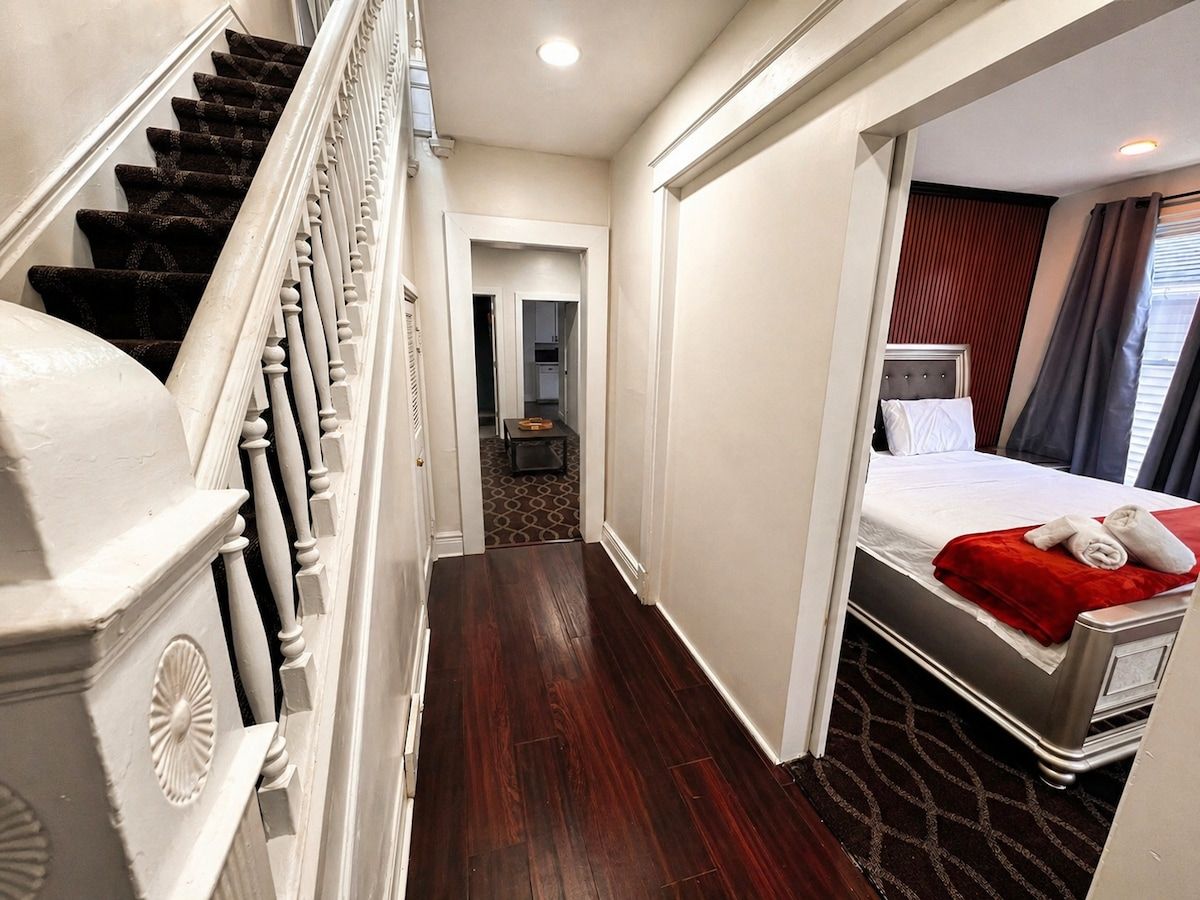 A hotel room with a bed, a red blanket, a dark wooden floor, a white wall, and a staircase.