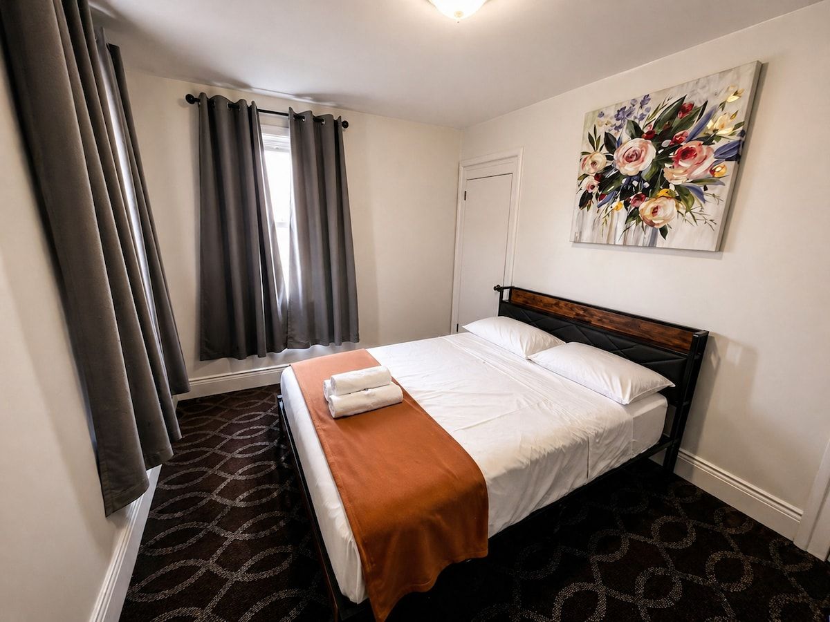 Hotellihuone, jossa on sänky, ikkuna verhoilla, taulu seinällä ja kuviollinen matto.
