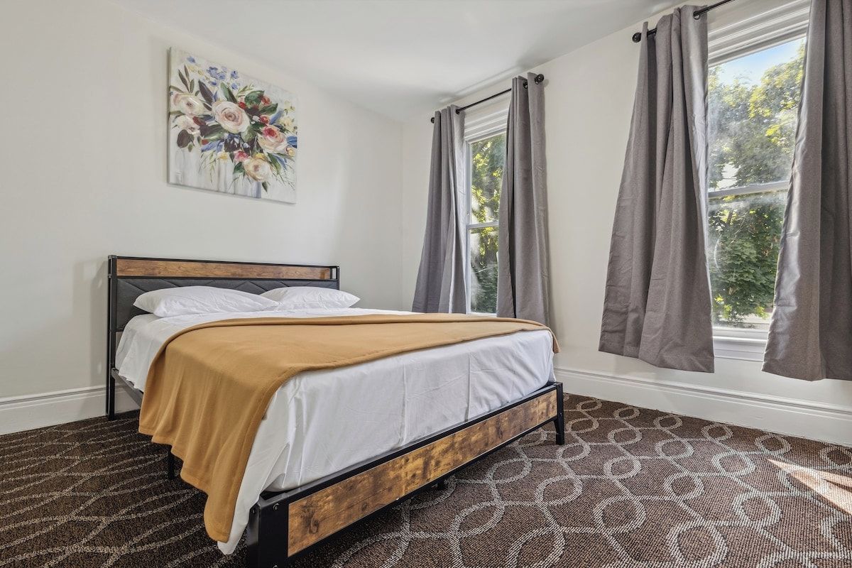 Un dormitorio con cama, ventana con cortinas, alfombra estampada y un cuadro floral en la pared.