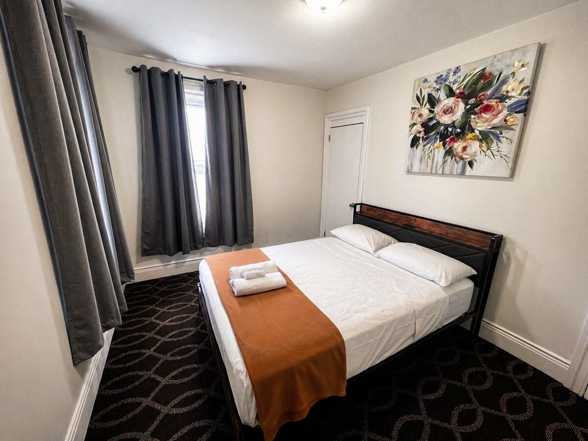 A hotel room with a bed, a window with curtains, a painting on the wall, and a patterned carpet.