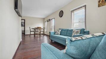 A living room with a blue sofa, a dining table with chairs, and a clock on the wall.
