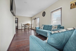 A living room with a blue sofa, a dining table with chairs, and a clock on the wall. - Cozy 2BR 1 Block to Falls Perfect Couple Getaway (Niagara Falls)