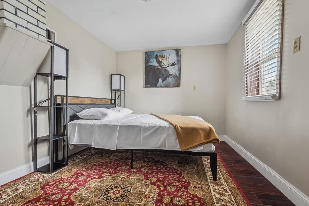 A bedroom with a bed, a red patterned rug, a wall-mounted shelf, and a painting of a moose.