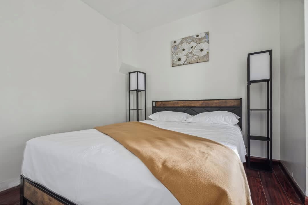 A single bed with a wooden headboard, white bedding, and a brown throw blanket. There is a wall-mounted artwork above the bed and a tall, narrow shelf on the right.
