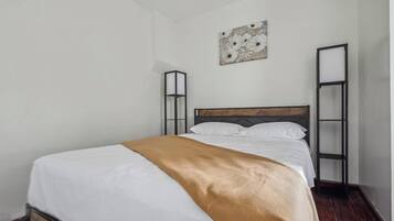 A single bed with a wooden headboard, white bedding, and a brown throw blanket. There is a wall-mounted artwork above the bed and a tall, narrow shelf on the right.