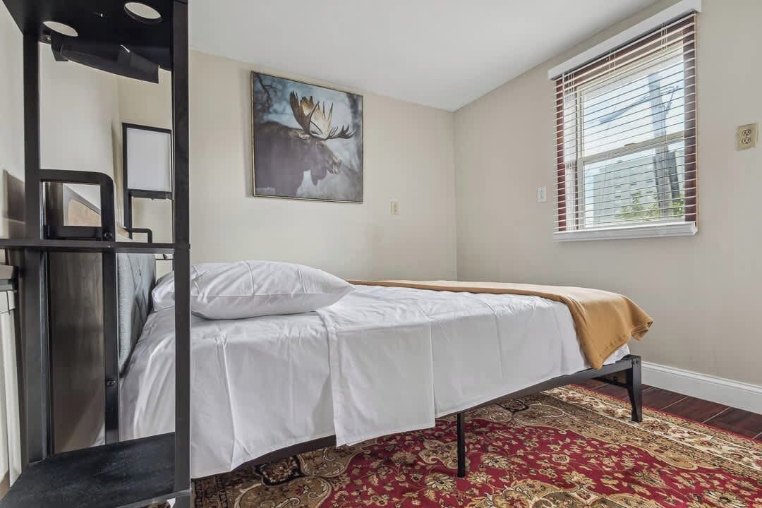 A single bed with a white mattress and a patterned red carpet, a painting of a moose on the wall, and a window with blinds.