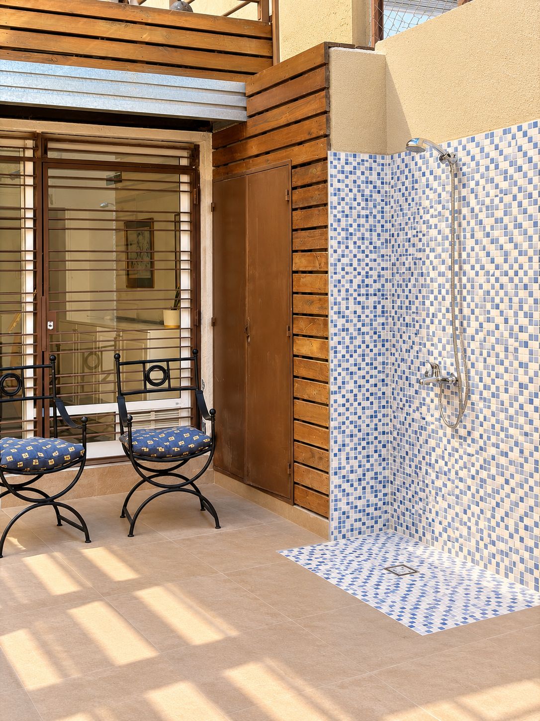 A covered outdoor area with a wooden deck, two chairs, and a shower area with blue and white mosaic tiles.