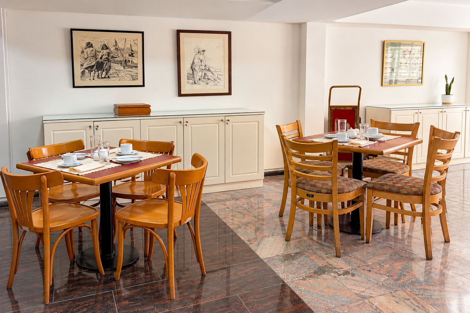A dining area with a table set for four, surrounded by wooden chairs. There are framed artworks on the walls, a glass cabinet with a red chair, and a potted plant on a table.