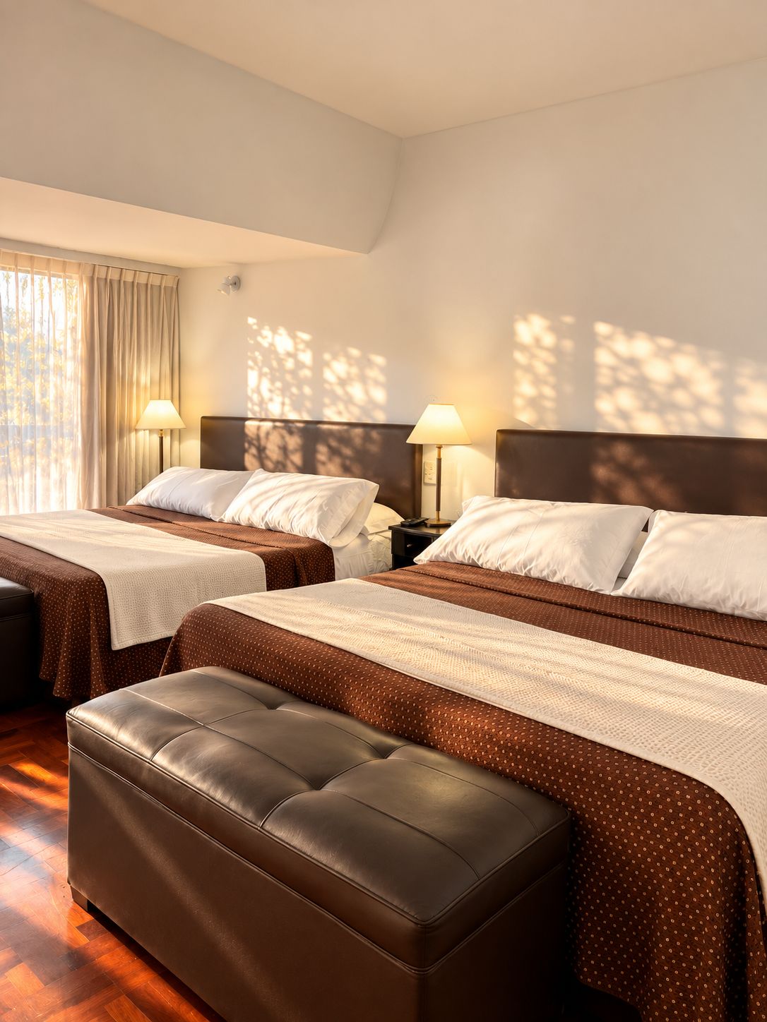 Une chambre d’hôtel avec deux lits, un pouf en cuir marron et une fenêtre avec des rideaux.