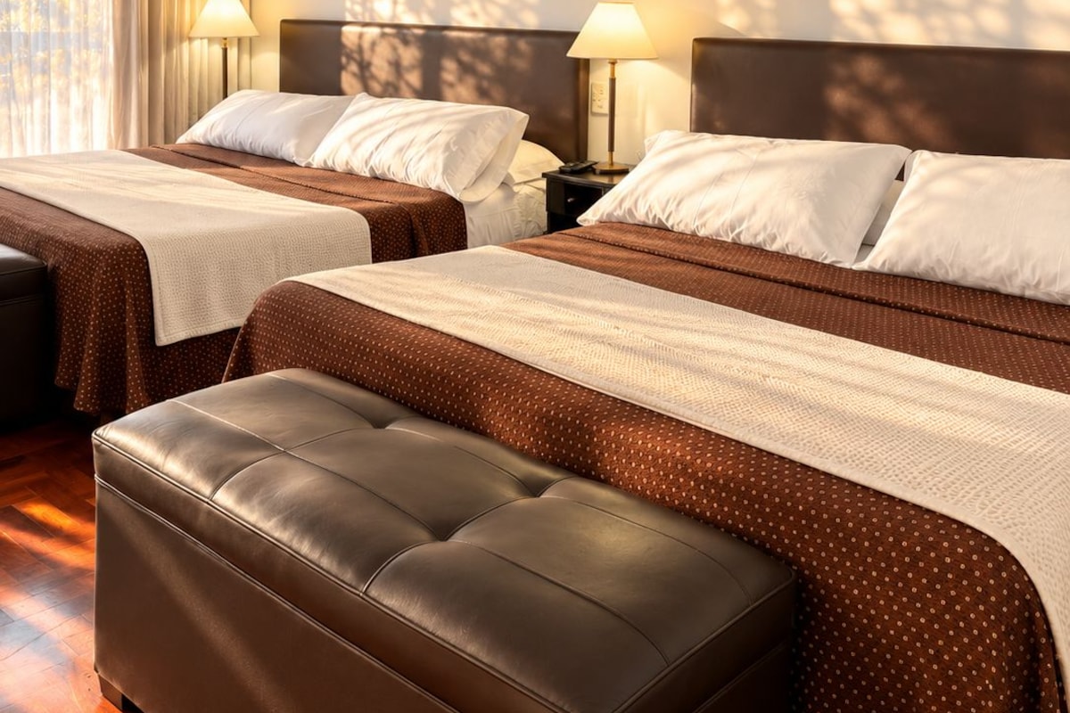 A hotel room with two beds, a brown leather ottoman, and a window with curtains.