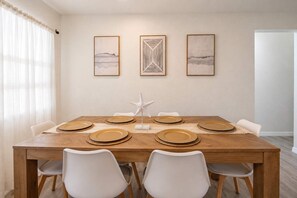 A dining room with a wooden table set for six, white chairs, and three framed artworks on the wall. - 5BR, Private Pool, Near Pier 60 by Golden Coast Rentals (Clearwater)