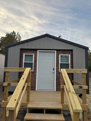 A small wooden building with a white door, two windows, and a ramp leading to the entrance. - Quaint Tiny Home with Kitchen (Columbia)