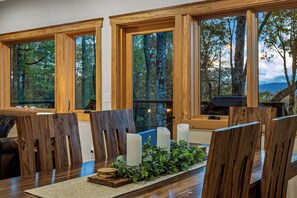 A dining area with wooden chairs and a table, large windows with a view of trees and mountains, and candles on the table. - Majestic Mountain View, Hot Tub, Outdoor Shower, Sauna & Game Room (Penrose)