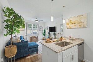 A modern kitchen with a granite countertop, stainless steel sink, and a dishwasher. There is a dining area with a blue sofa and a plant. - Grand Boho Suite / Queen Bed-Pool-Gym Near Airpot (Jacksonville)