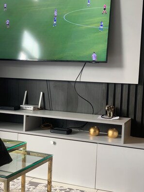 A flat-screen TV mounted on a wall, with a gaming console and decorative items on a shelf below. - Charming apartment in beautiful Accra with cool AC and gym access (Accra)