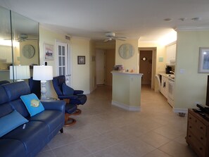 A living room with a blue sofa, a small island with a clock, and a kitchen area with appliances. - 2-bedroom beachfront condo in Marco Island with fitness room,pool,picketball (Marco Island)