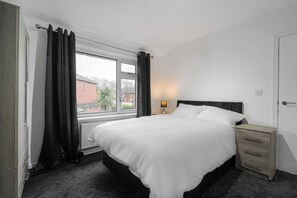 A bedroom with a bed, a bedside table, a lamp, and a window with curtains. - Refurbished Farnworth Home Comfort and Convenience Await for Holiday Makers (Bolton)
