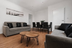 A modern living room with a grey sofa, a round wooden coffee table, and a dining area with black chairs and a table. - Refurbished Farnworth Home Comfort and Convenience Await for Holiday Makers (Bolton)