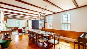 A spacious dining area with wooden flooring, a long table set for a meal, and large windows providing natural light. - Rosegate Cottage | Panoramic Harbor Views + Walk Downtown (Boothbay Harbor)