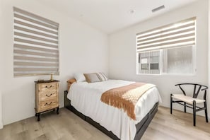 A bedroom with a bed, a wooden dresser, a chair, and two windows with blinds. - Modern 3-bedroom Coastal Retreat w/ Ocean & Mountain Views (Costa Mesa)