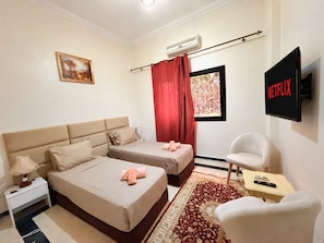 Ein Hotelzimmer mit zwei Betten, einem Fernseher, einem Sessel und einem fensterbepflanzten Fenster.