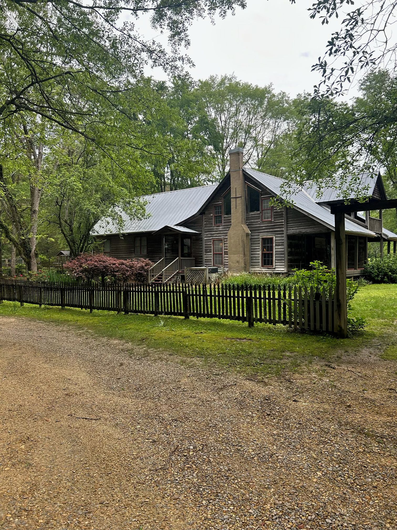 Une maison rustique à deux étages, avec un toit en métal, entourée d’une clôture en bois et d’une végétation luxuriante.