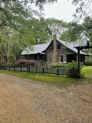 A rustic two-story house with a metal roof, surrounded by a wooden fence and lush greenery.