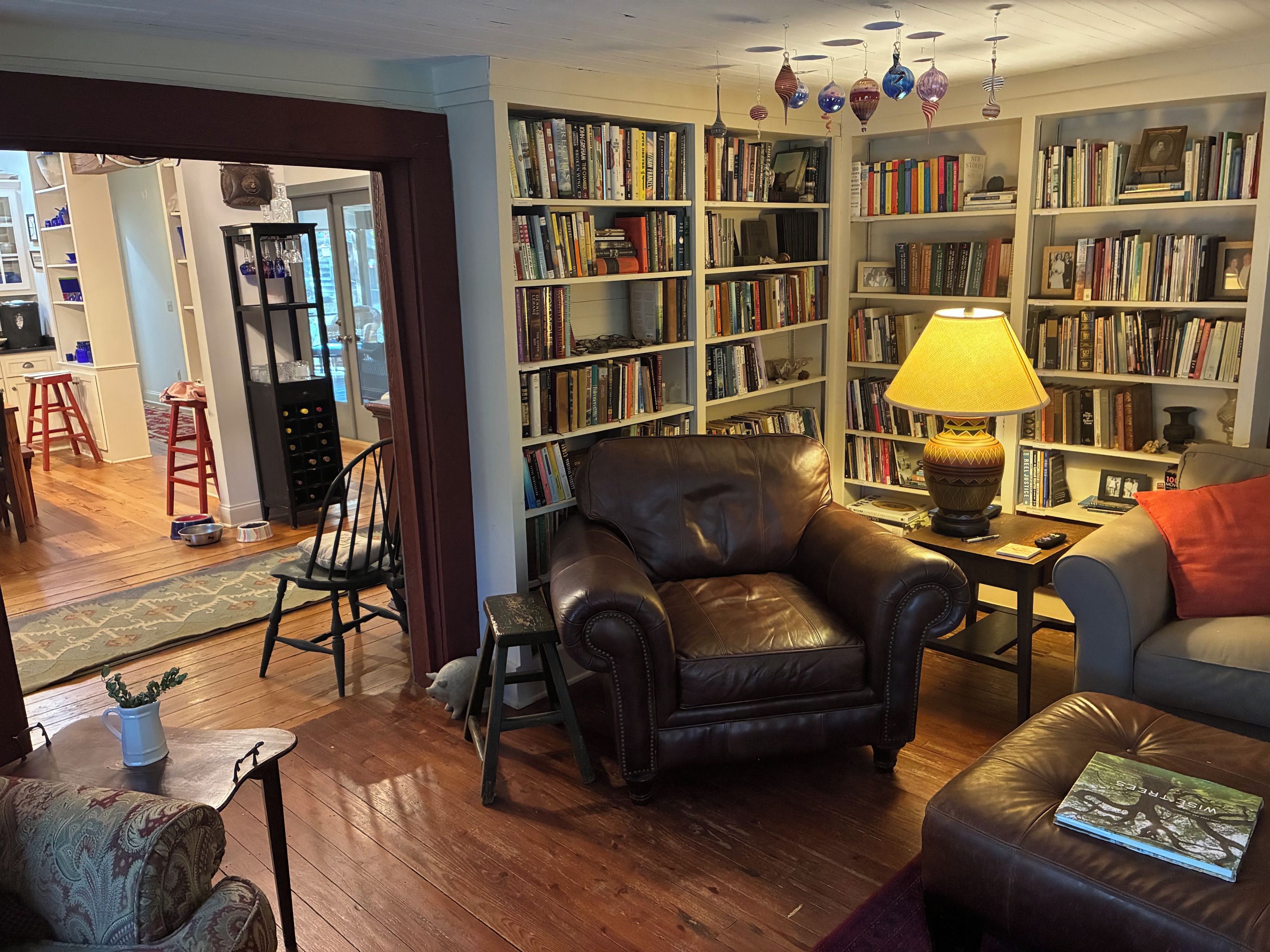 Un salon chaleureux avec un fauteuil en cuir, une bibliothèque remplie de livres, une petite table avec une lampe et un canapé gris.