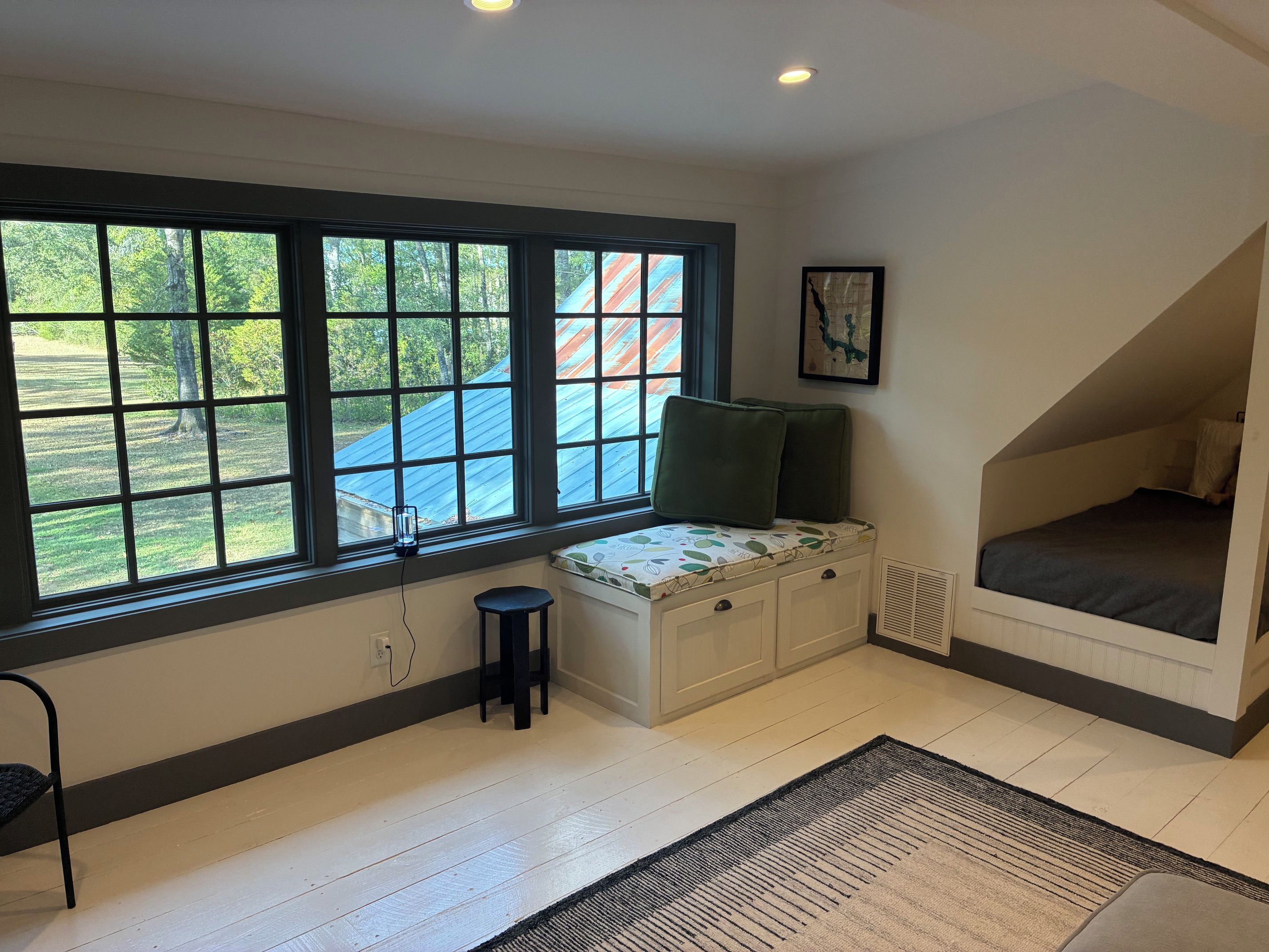 Une pièce avec une grande fenêtre, un banc avec des coussins, un petit tabouret et un lit situé dans un espace en mezzanine.