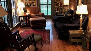 A cozy living room with wooden flooring, a leather armchair, a sofa, a rocking chair, and a bookshelf filled with books.