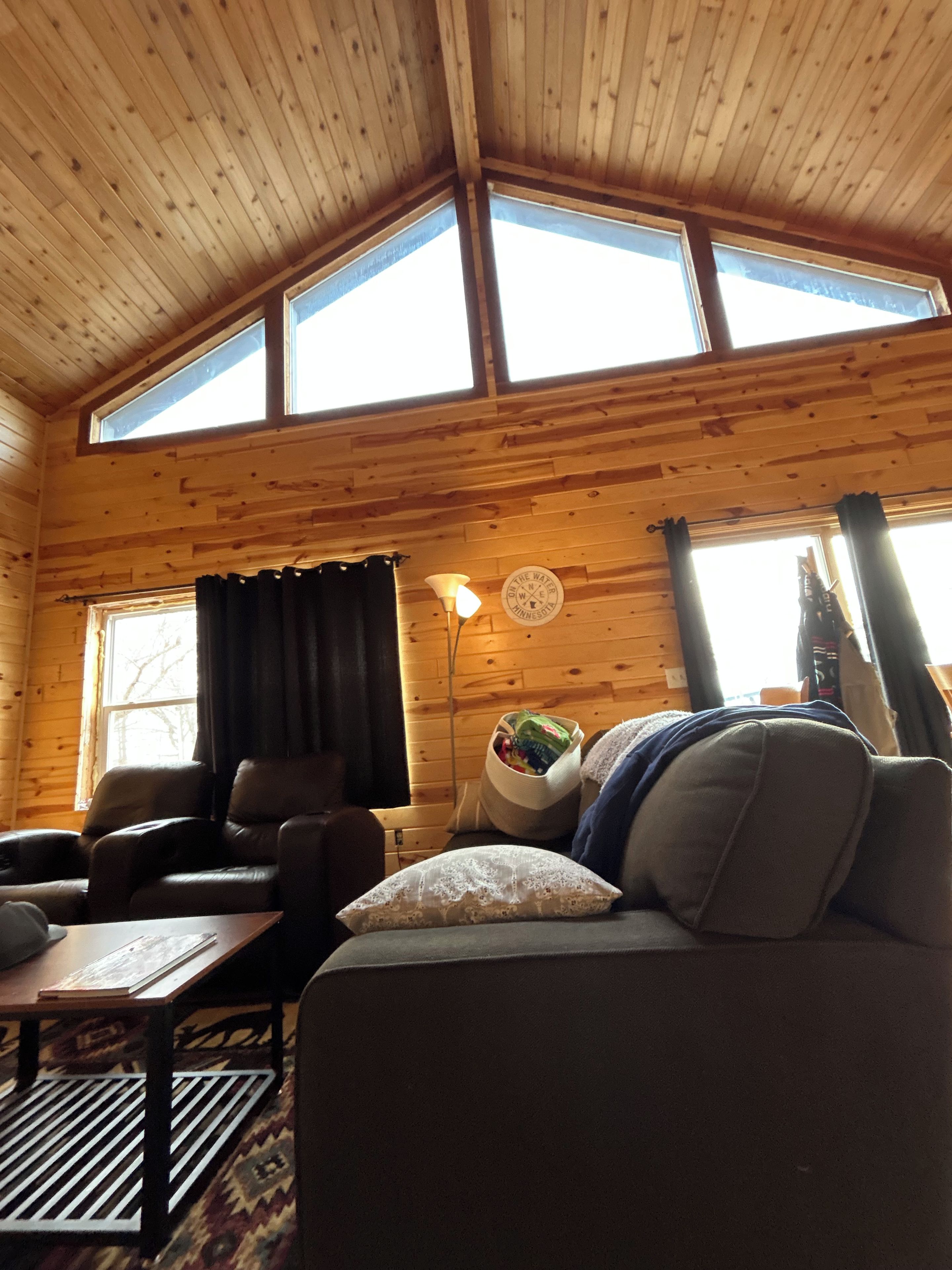 A cozy cabin interior with a wooden ceiling, a sofa, a table, and a lamp.