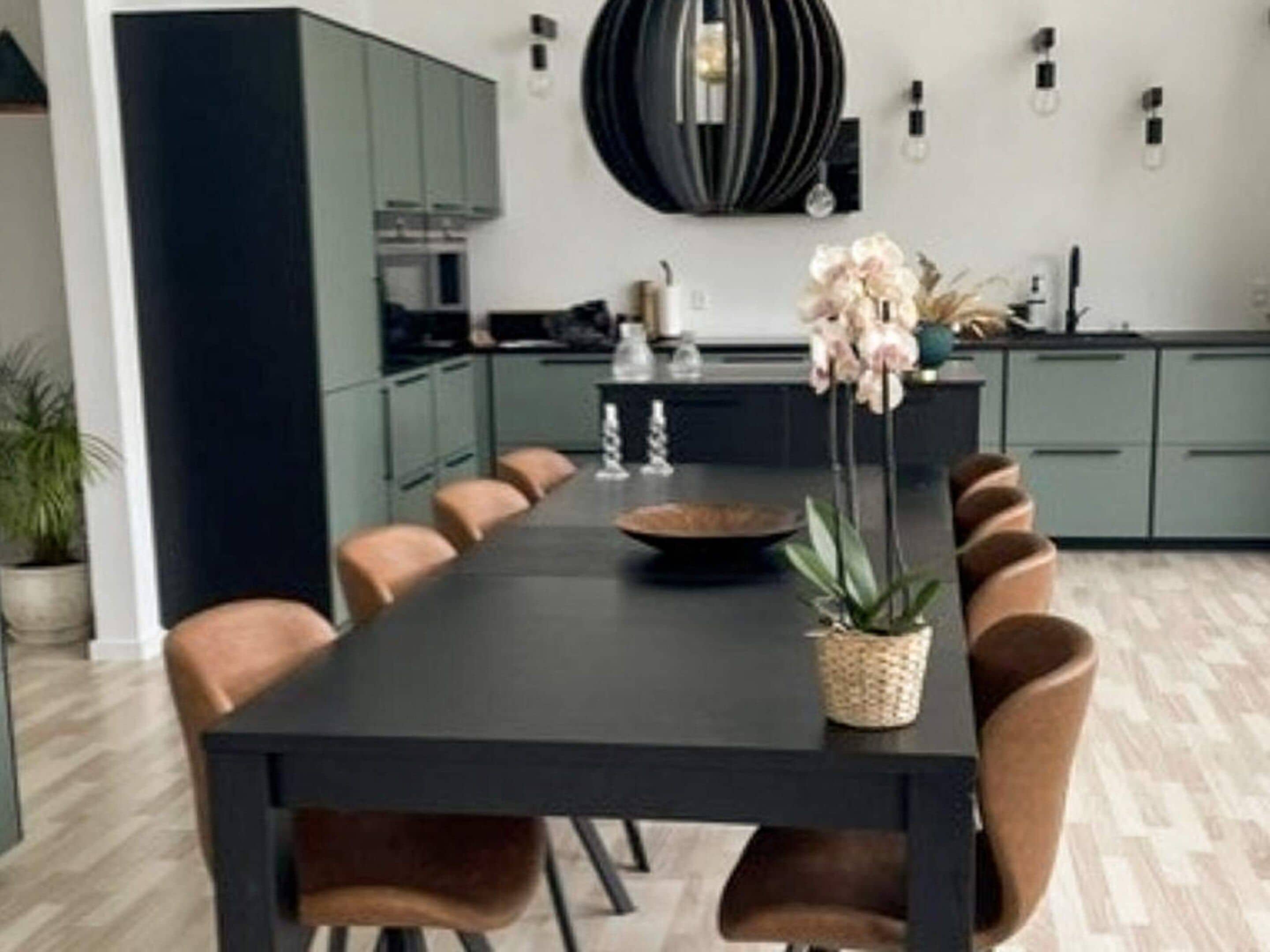 Un comedor moderno con una mesa larga, múltiples sillas, un jarrón central con flores y una cocina al fondo.