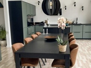 A modern dining area with a long table, multiple chairs, a central vase with flowers, and a kitchen in the background. - 4 Star Holiday Home in Gilleleje (Gilleleje)