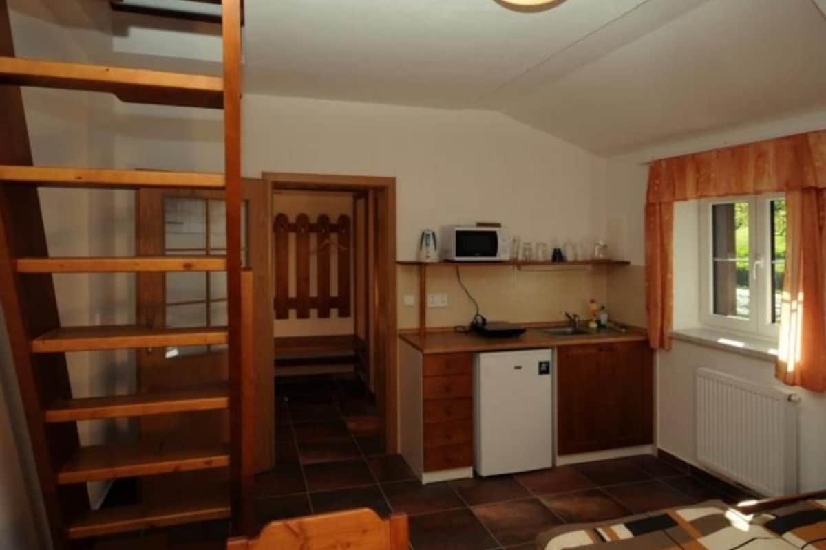 Una camera compatta con un letto, una scrivania con computer, un piccolo frigorifero e una finestra con tende.