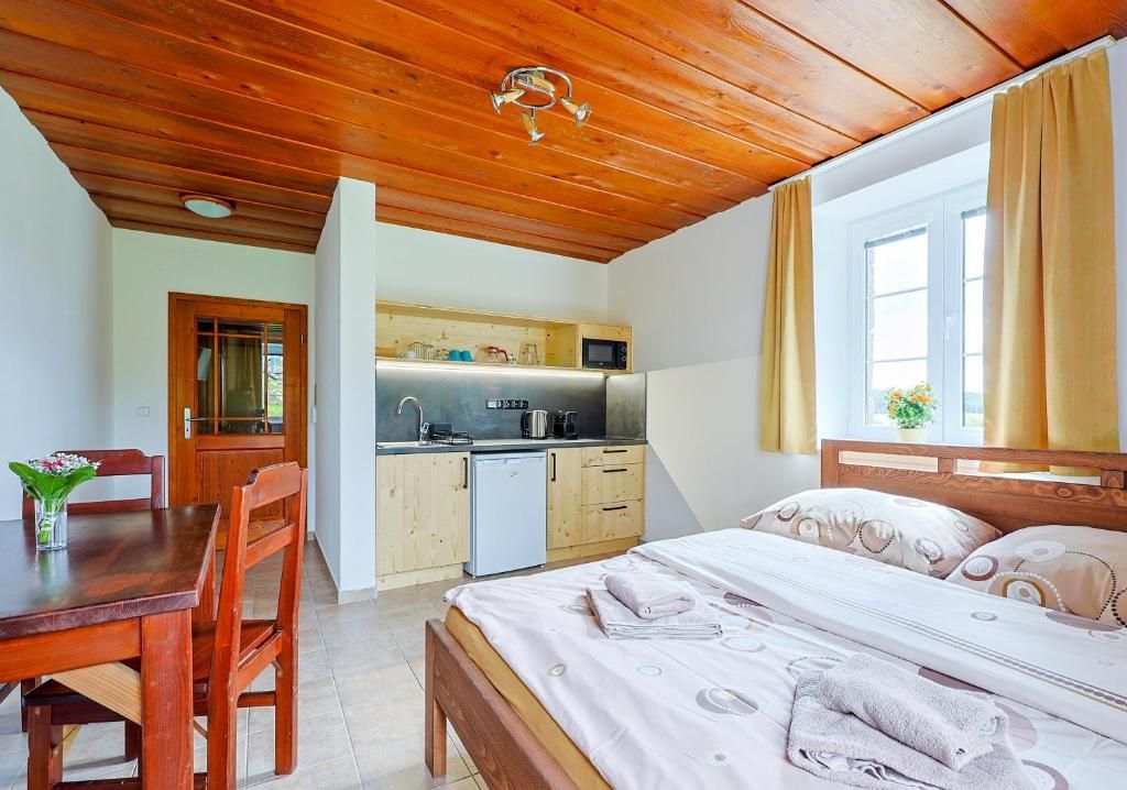Una stanza con soffitto in legno, un letto con copriletto bianco, un tavolo da pranzo in legno con sedie, un angolo cottura con forno a microonde e una finestra con tende.