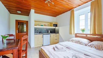 Una stanza con soffitto in legno, un letto con copriletto bianco, un tavolo da pranzo in legno con sedie, un angolo cottura con forno a microonde e una finestra con tende.
