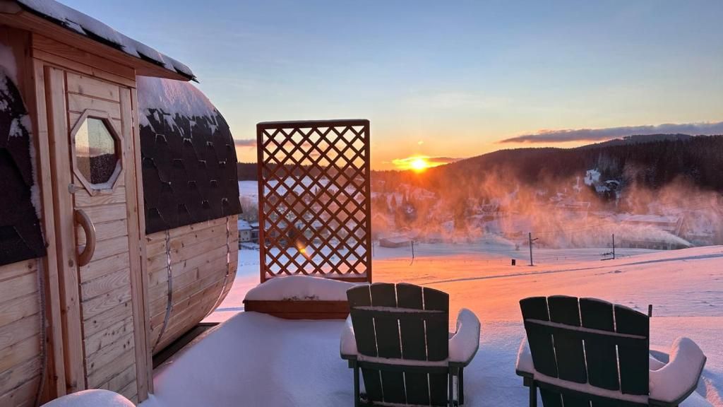 A wooden cabin with a snow-covered roof, a lattice screen, and two Adirondack chairs facing a snowy landscape with a sunset.